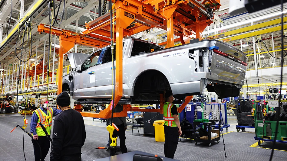 Two autoworkers stand beside the frame of an elevated all-electric pickup truck body on the assembly line, while another works beneath it.