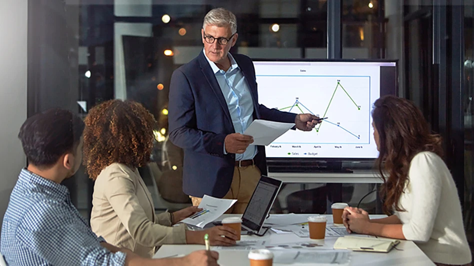 A man points to a graph on a screen while others watch from a table.