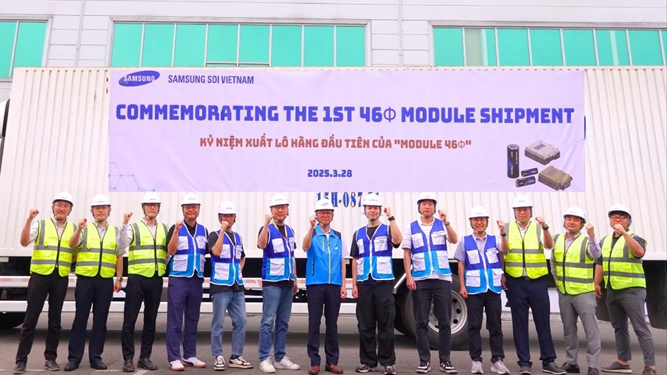 Several people stand in front of a building with a banner on it reading Commemorating the 1st 46 Module Shipment